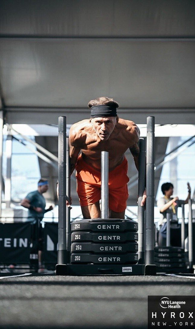 A client pushing a weighted sled at a HYROX race under dramatic side light